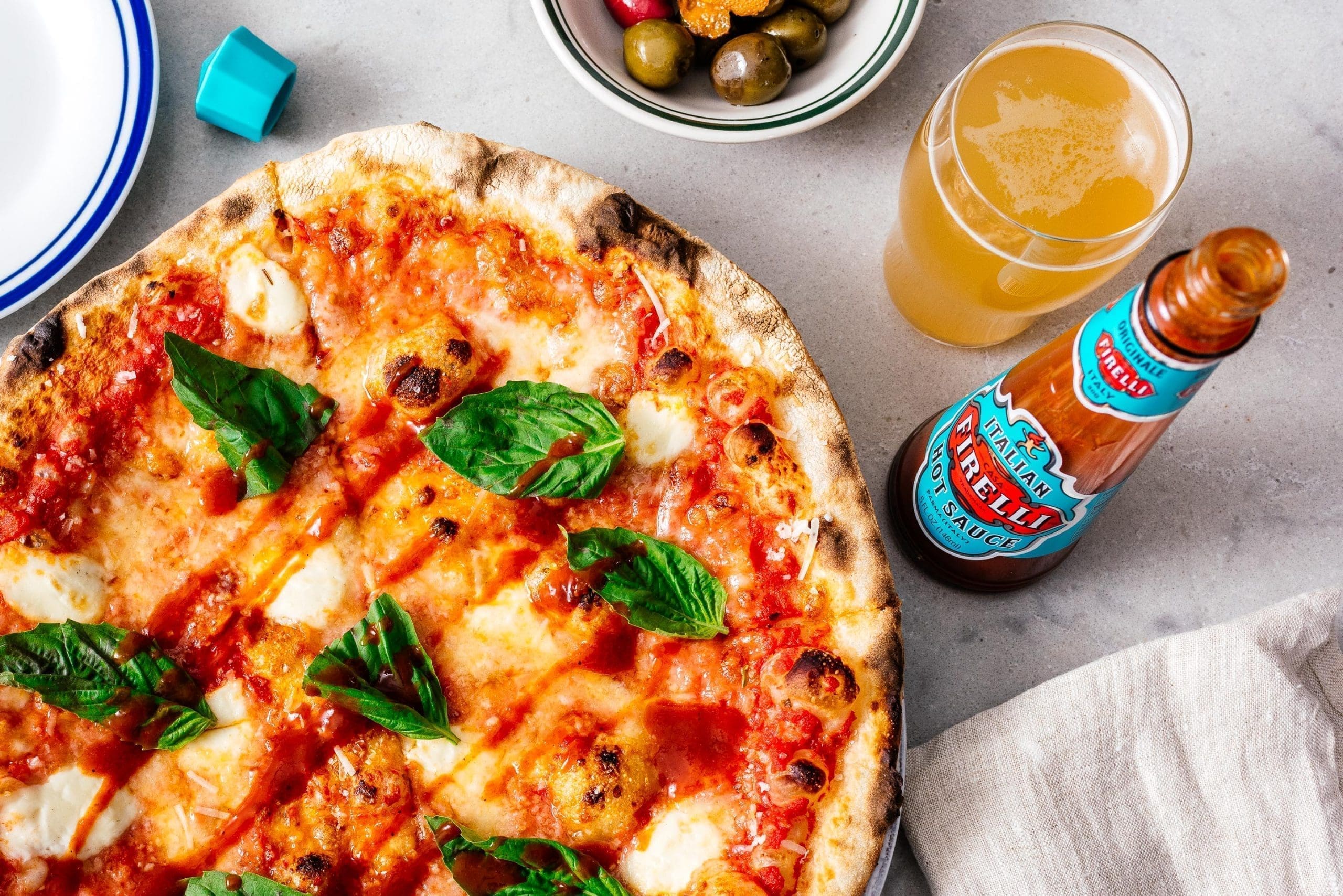 a bottle of firelli hot sauce next to a pizza and a beer